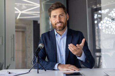 Profesyonel bir erkek iş adamı tek eliyle podyumun arkasından izleyiciye güven içinde seslenir. Doğal ışıklandırma ile modern ofis arka planı.