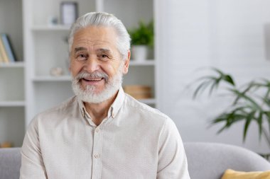 Gülümseyen, beyaz sakallı, modern bir oturma odasında oturan, mutluluk ve sağlık saçan son sınıf öğrencisi..