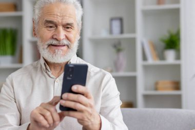 Beyaz gömlekli, gri saçlı ve akıllı telefon kullanan yaşlı ve gülümseyen bir büyükbabanın yakın plan fotoğrafı..