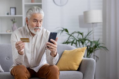 Beyaz saçlı ve sakallı son sınıf öğrencisi evdeki kanepede oturuyor elinde bir banka kartı tutuyor ve endişeli bir şekilde cep telefonu ekranına bakıyor..