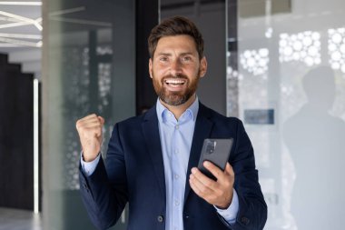 Sakallı neşeli bir işadamı, modern ofis ortamında akıllı bir telefonu elinde tutarak mutluluğu ve başarıyı kutluyor..