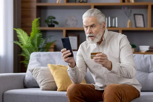 stock image A gray-haired shocked senior man is at home, holding a credit card and worriedly looking at the mobile phone screen.