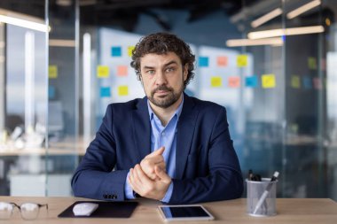 Kendine güvenen mavi takım elbiseli işadamı modern ofiste dijital tablet ve ofis malzemeleriyle masada oturuyor. Arka planda cam duvarda yapışkan notlar olan profesyonel şirket ayarları.