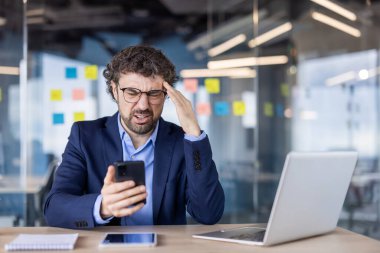 Stresli iş adamı, modern ofiste hayal kırıklığına uğramış bir ifadeyle telefonu elinde tutuyor. İş profesyonelleri iş zorluklarıyla uğraşıyor. Masada dizüstü bilgisayar ve not defteri, cam duvarda renkli notlar..