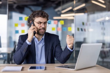 Modern ofis ortamında iş adamı telefon görüşmesi sırasında hayal kırıklığı gösteriyor. Takım elbiseli bir adam dizüstü bilgisayarda çalışırken öfkesini ifade ediyor. Dijital tablet ve dizüstü bilgisayarla..