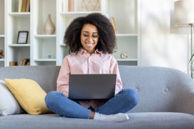 Evde dizüstü bilgisayarla çalışan, oturma odasında kanepede oturan gülümseyen bir kadın. Uzaktan çalışmaktan zevk alan rahat genç bir profesyonel, günlük kıyafetler. Konforlu ev ortamı modern yaşam tarzı dengesini gösteriyor.