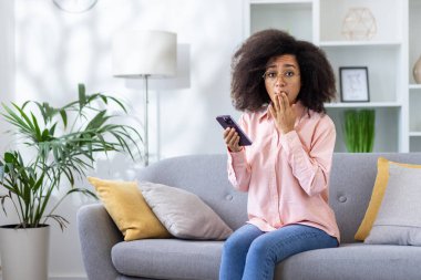 Afro saçlı, gözlüklü ve pembe tişörtlü bir kadın. Kanepede oturmuş elinde akıllı bir telefonla şaşırmış bir ifadeyle oturuyor. Arka planda bitkiler olan modern oturma odası..