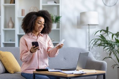 Akıllı telefon ve dizüstü bilgisayarı internet üzerinden ödeme yapmak için kullanan gülümseyen bir kadın. Modern oturma odasındaki dijital bankacılık, uzaktan çalışma ve finansal yönetim kavramı.