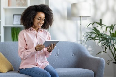 Parlak oturma odasındaki gri koltukta oturan genç bir kadın dijital tablet kullanıyor ve kulaklıkla müzik dinliyor. Modern dekorlu ve doğal ışıklandırmalı rahat bir ev ortamı..