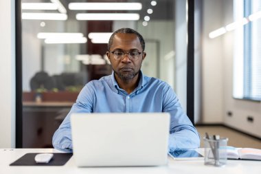 Modern ofiste dizüstü bilgisayarda çalışan bir iş adamı. Konsantrasyon ve verimliliği teşvik eden profesyonel bir ortam. Çalışma ortamını geliştiren çağdaş tasarım elementlerine sahip iyi ışıklandırılmış bir oda.