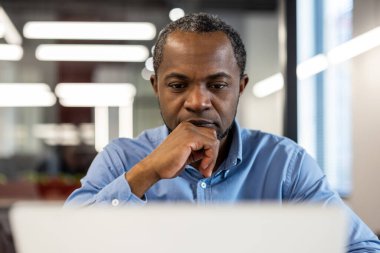 Modern ofis ortamında dizüstü bilgisayar kullanan profesyonel iş adamı. Ciddi ifade derin düşünceyi yansıtır. İş, teknoloji ve verimliliği vurgulayan konular için ideal