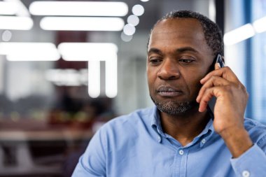 Modern ofiste telefon görüşmesi yapan profesyonel bir iş adamı. Adam konuşmaya odaklanmış. Bulanık geçmişi olan, ofis ortamını ve teknoloji kullanımını vurgulayan parlak, çağdaş çalışma alanı.