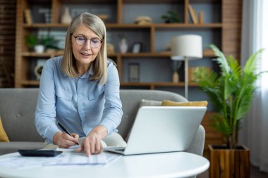 Evde dizüstü bilgisayarla çalışan, belgeleri inceleyen ve modern oturma odasında hesap makinesi kullanan kıdemli bir kadın. Arka planda masa ve kitaplık ile ev ofisi kurulumu. Uzak çalışma verimliliği kavramı.