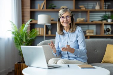 Üst düzey bir kadın, müşterileriyle evdeki bilgisayarından iletişim kuruyor, gülümsüyor ve kulaklık kullanıyor. Masanın üzerinde bitki, kitap ve not defterleriyle rahat bir ofis düzeni var.