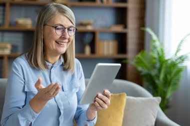 Oturma odasında videolu görüşme için tablet kullanarak gülümseyen son sınıf kadını. İletişim, teknoloji, mutluluk, aile kavramı ve uzaktan bağlı kalmak..