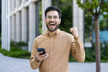 Telefonu elinde tutan heyecanlı genç adam, modern ofis binasının dışındaki başarıyı kutluyor. Günlük kıyafetler giymek, neşe ve başarı hissetmek, şehir ortamında açık havada.