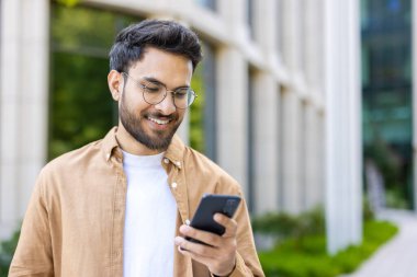 Modern binanın yakınında akıllı telefon kullanırken gülümseyen gözlüklü ve günlük kıyafetler giyen bir adam. Güneşli bir günde açık havada iletişimi ve teknolojiyi seven kişi