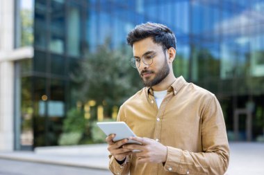 Genç adam dijital tablet kullanarak modern cam ofis binasının önünde duruyor. İşle ilgili görevlere odaklanmak, kentsel ortamdaki mesleki ve teknolojik hayat tarzını yansıtmak.