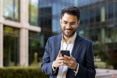 Modern cam ofis binasının dışında akıllı telefon kullanırken gülümseyen takım elbiseli kendine güvenen genç iş adamı. İş, iletişim, teknoloji, güven ve mesleki yaşam tarzı kavramı.