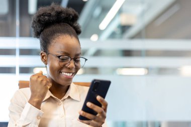 Ofisteki mutlu kadın modern ortamda heyecanlı akıllı telefon kullanıyor. Yumruk pompasıyla başarıyı kutlamak, gözlük takmak ve beyaz gömlek giymek, başarı anının tadını çıkarmak.