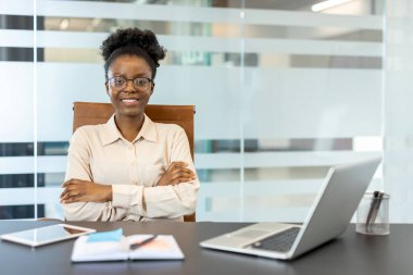Modern ofiste masasında oturan kendine güvenen bir iş kadını, kollarını kavuşturmuş, gülümsüyor. Masadaki dizüstü bilgisayar, belgeler ve tablet verimliliği ve profesyonelliği gösteriyor.