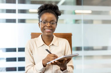 Modern ofisteki video görüşmesi sırasında elinde kalem ve defter tutan kendine güvenen bir iş kadını. Gülümseyen profesyonel notlar alıyor, bağlılığı ve hazırlığı gösteriyor.