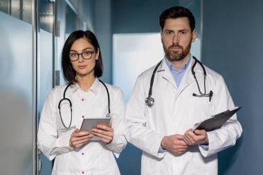 Hastane koridorunda duran iki profesyonel doktor. Kadın doktor elinde tablet tutuyor. Elinde dosya tutan bir erkek doktor. Beyaz önlük ve steteskop giyen resmi giyinmiş tıp uzmanları..