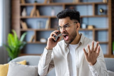 Öfkeli adam evde telefonda bağırıyor. Hayal kırıklığı ve stresi dile getiriyor. Genç yetişkin gözlüklü hararetli bir konuşma yapıyor modern oturma odasında yoğun duygular sergiliyor..