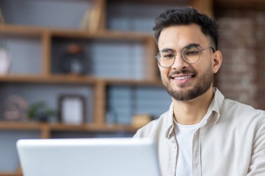 Modern ofis bilgisayarında çalışırken gülümseyen gözlüklü genç bir adam. Günlük kıyafetler, arka planda kitaplıklar. Uzaktan çalışma, üretkenlik, ev ofisi ve teknoloji kavramı.