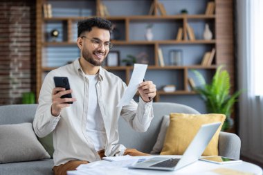 Akıllı telefon ve dizüstü bilgisayarı kullanarak evdeki rahat oturma odasının finansmanını sağlamak için gülümseyen genç adam. Faturalar ve evrak işleri konusunda hevesli. İçişleri Bakanlığı kavramı, mali yönetim