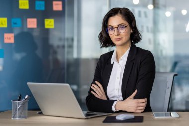 Modern ofis ortamında dizüstü bilgisayarda çalışan kendine güvenen bir iş kadını. Yapışkan notları ve masa aksesuarları olan profesyonel çalışma alanı. İş, ofis ve mesleki temalar için ideal.
