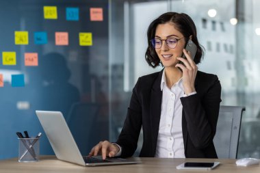 Modern ofis ortamında kendine güvenen bir iş kadını. Dizüstü bilgisayarla uğraşırken, cam duvardaki renkli yapışkan notlarla meşgul olmuş.