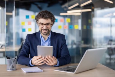 Modern ofis ortamında dijital tablet kullanan bir iş adamı. Modern dekor, yapışkan notlar, dizüstü bilgisayar ve not defteri ile çevrili çalışma faaliyetleri, verimliliği taşıma