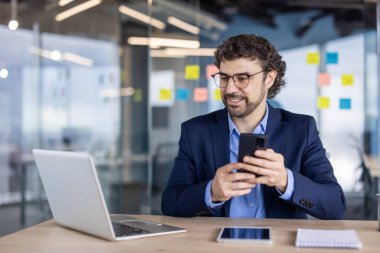 Ofisteki kendine güvenen iş adamı akıllı telefon ve dizüstü bilgisayarla çalışıyor. Dijital cihazlı profesyonel çalışma ortamı, cam üzerine yapışkan notlar ve modern tasarım. Kurumsal ayar tanıtımı