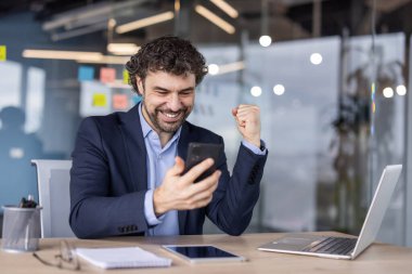 Takım elbiseli mutlu işadamı modern ofisteki akıllı telefondan başarıyı kutluyor. Masada dizüstü bilgisayar, not defteri ve gözlüklerle profesyonelce heyecan ve neşe ifade ediyor. İş başarısı kavramı