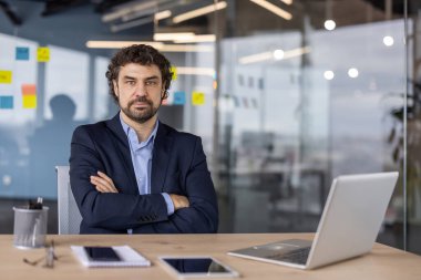 Modern ofiste kolları bağlı oturan kendinden emin bir iş adamı. Masasında dizüstü bilgisayarı olan profesyonel bir kıyafet. İş, liderlik ve ofis işi kavramı.