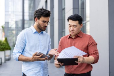 Modern ofis binasının önünde duran iki işadamı, belgeler ve tabletleri incelerken sohbet ediyorlar. İş ortamında takım çalışması, işbirliği ve iletişim kavramı.