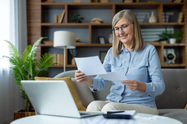 Üst düzey kadın evde bilgisayarla çalışırken kanepeye oturmuş belgeleri inceliyor. Konforlu oturma odası ortamında odaklanmış ve içerikli görünüyor, uzak iş veya kişisel görevlerle meşgul