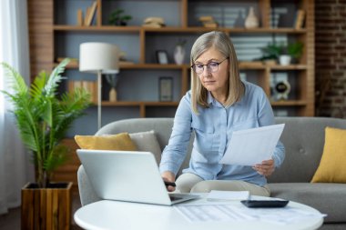 Evde dizüstü bilgisayarla çalışan kıdemli bir kadın. Belgeleri gözden geçiriyor ve görevlerine odaklanıyor. Uzaktan çalışma, ev ofisi, üretkenlik ve kıdemli profesyonellik kavramları.