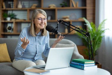 Gülümseyen son sınıf öğrencisi kadın podcast 'i kaydediyor, kanepede dizüstü bilgisayarı ve mikrofonuyla oturuyor. Çevrimiçi iletişim, bilgi paylaşımı ve izleyicilerle iletişim kurma. Uzak çalışma kavramı ve