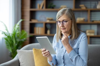 Modern oturma odasında gözlüklü, tablette yazan son sınıf kadını. Konsantrasyon ve merak gösteren cihazla çok ilgili görünüyor..