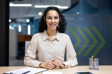 Gülümseyen kadın müşteri hizmetleri temsilcisi modern ofis çalışma alanında kulaklık takıyor. Kendine güvenen profesyonel yardım, belgeler ve tablet masası kurumsal ortamı ile çalışmak.