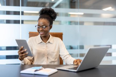 Modern ofiste masa başında dizüstü bilgisayar ve tablet kullanan bir iş kadını. Profesyonel gözlük takıyor, iş programını dengelerken gülümsüyor. Çoklu görev, üretkenlik kavramı