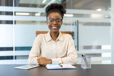 Modern ofiste masa başında otururken gülümseyen kendine güvenen bir iş kadını. Not defteri ve tabletle çalışan profesyonel bir kadın. Video ve ofis görevlerine hazır..
