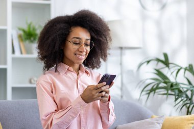 Modern oturma odasında akıllı telefon kullanan gülümseyen bir kadın. Kapalı alan bitkileri ve doğal ışık ile aydınlık ve neşeli bir atmosfer ev ortamını rahatlatıyor..