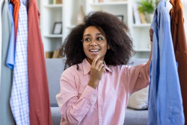 Kıvırcık saçlı ve gözlüklü bir kadın gardıroptan kıyafet seçiyor, kıyafet seçimlerini düşünürken gülümsüyor. Konforlu ev ortamı, moda ve tarz kavramları