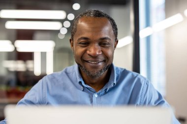 Modern ofiste dizüstü bilgisayarla çalışan mavi gömlekli gülen bir ofis çalışanı. Verimliliği ve pozitifliği vurgulayan parlak, profesyonel ortam.