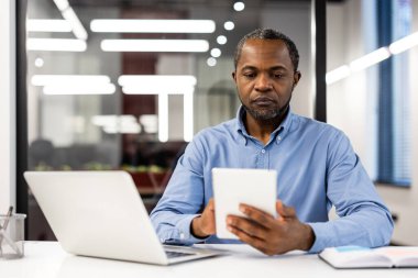 Modern ofis ortamında tablet kullanan bir ofis çalışanı. Adam masa başında dizüstü bilgisayar ve belgelerle çalışmaya odaklanmış. İş, teknoloji, üretkenlik ve iş hayatı kavramı.