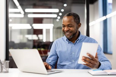 Modern ofis ortamında dizüstü bilgisayar ve tablet üzerinde çalışan mutlu iş adamı. Mavi gömlekli, çoklu görev yapan ve iş günlerinde teknolojiyle uğraşan bir profesyonel. Parlak ofis cam duvarları