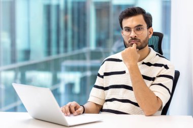 Gözlüklü ve çizgili tişörtlü dalgın bir ofis çalışanı modern çalışma ortamında dizüstü bilgisayarla masada oturuyor. Çalışma stratejilerini ve kararlarını yansıtıyor. Cam duvarlı çağdaş ofis ortamı.